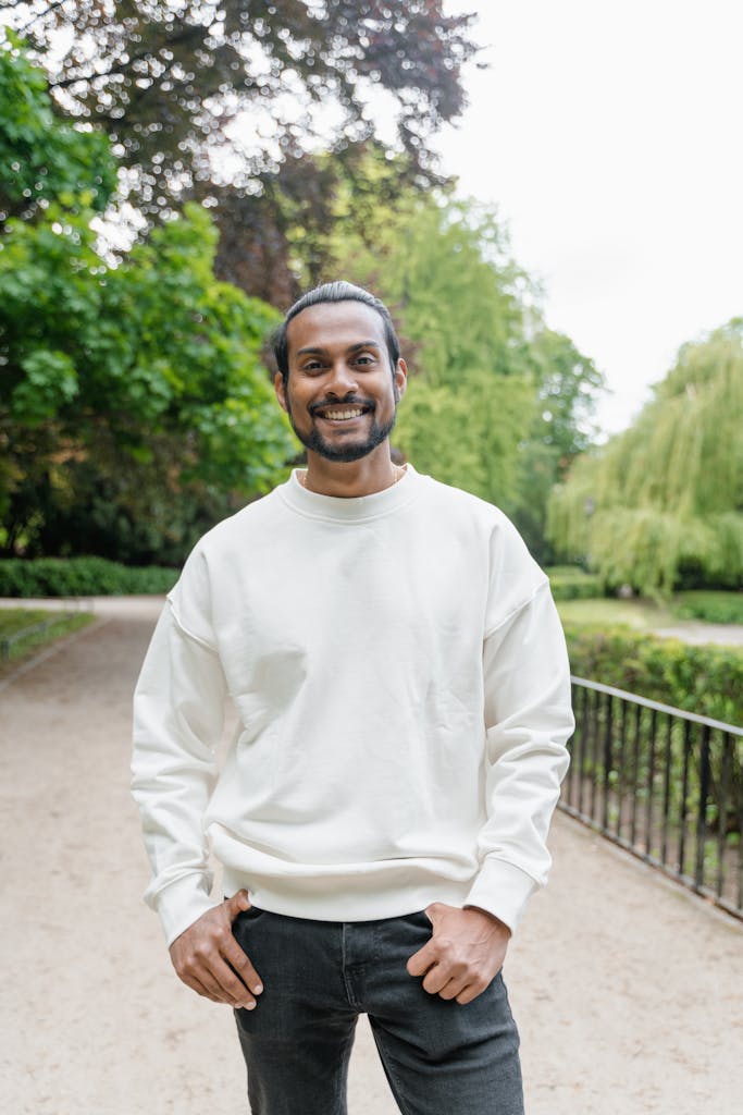 A cheerful man in a white sweater posing outdoors in a lush green park.